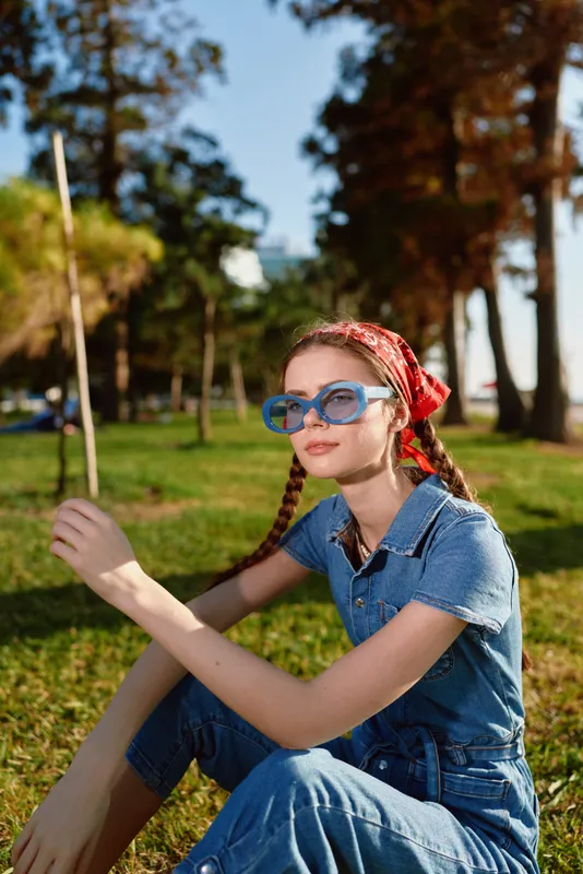 young woman with braids wearing blue glasses and denim outfit sitting outdoors on grass with green trees and warm sunlight in background
