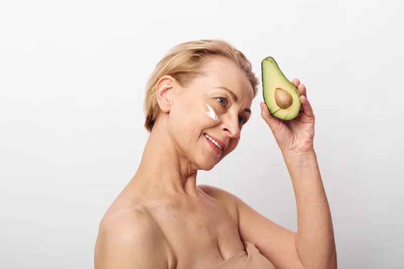 Mature woman with short blonde hair holds half of avocado near face, smiling gently. Cream patches under eyes suggest skincare routine with natural ingredients, isolated on white background.