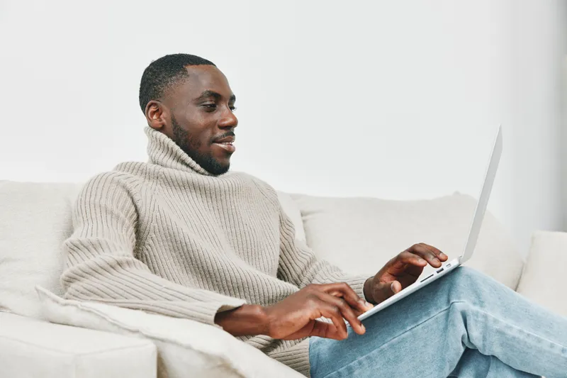 A young Black man in a cozy turtleneck sweater, joyfully working on his laptop at home, emphasizing productivity and comfort in a modern living space