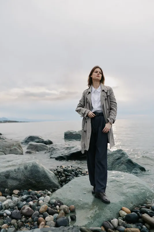 Woman in long coat and dark pants stands on rocky shore by the sea under cloudy sky. Fashion outdoor portrait with natural scenery and casual style. Concept of autumn and confidence.