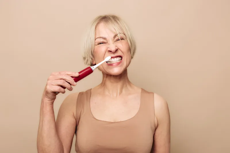 Blonde woman in beige tank top brushing teeth with electric toothbrush, making playful grimace. Oral hygiene, dental care, personal routine, health and wellness concept on neutral background.