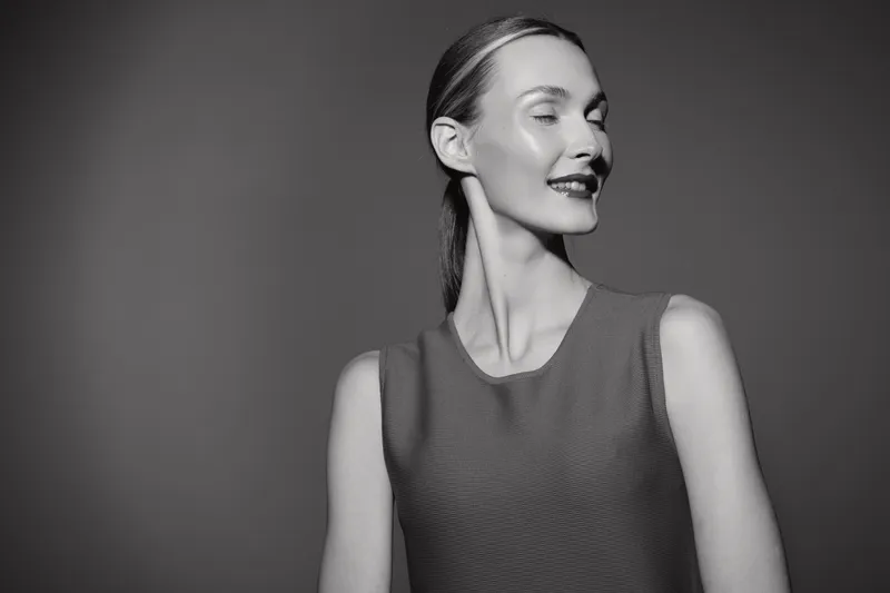 Smiling young Caucasian woman with fair skin poses with closed eyes and relaxed expression in studio with dark background. Concept of beauty and confidence.