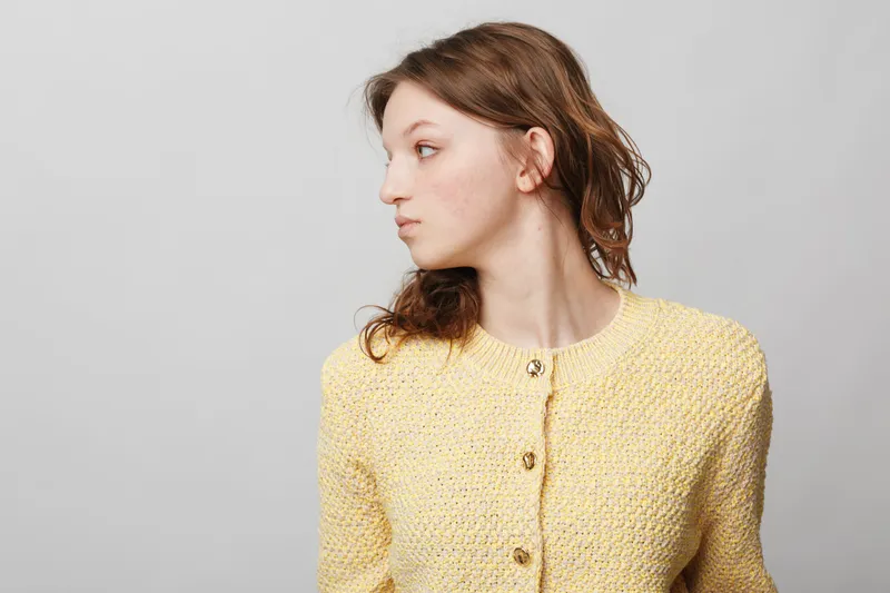 Young woman in yellow knitted sweater looks sideways with neutral expression on grey background. Concept of casual fashion, portrait, and natural beauty in studio lighting.