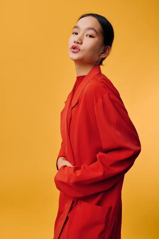 Young woman in red oversized jacket stands against yellow background looking sideways with relaxed expression casual fashion portrait stylish outfit and confident mood.
