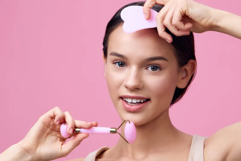 Young woman using a facial roller and guasha tool on her face with a soft pink background, representing self care and beauty routine for healthy skin