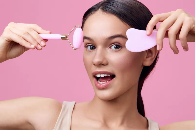 Smiling young woman demonstrating skincare tools, including a pink jade roller and heart shaped massager, against a vibrant pink background