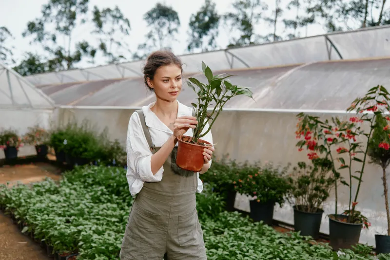 Young woman caring for a potted plant in a greenhouse, showcasing passion for gardening and sustainable living, with greenery in the background