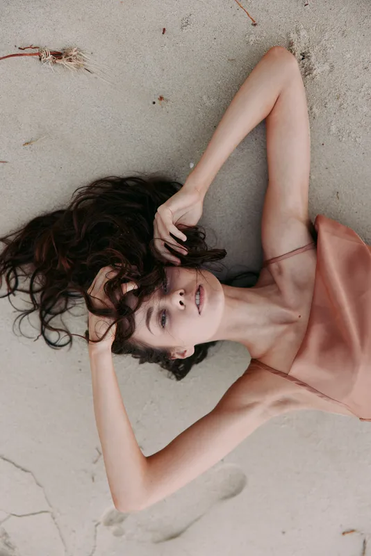 Portrait of a woman lying on sandy beach in a pink satin dress, with tousled hair and a tranquil expression, capturing soft light and a serene summer mood.