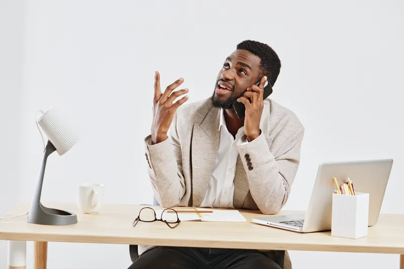 Male business professional talking on mobile phone while sitting at desk with laptop and office supplies in a bright modern workspace. People lifestyle concept, communication, technology, work from ho
