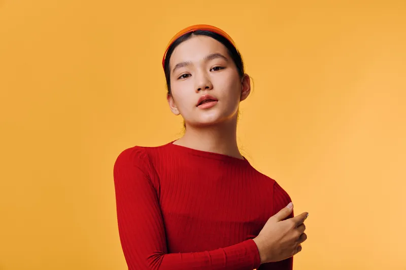 Young woman wearing red sweater and headband posing with hand on shoulder against orange background for casual fashion portrait and style concept.