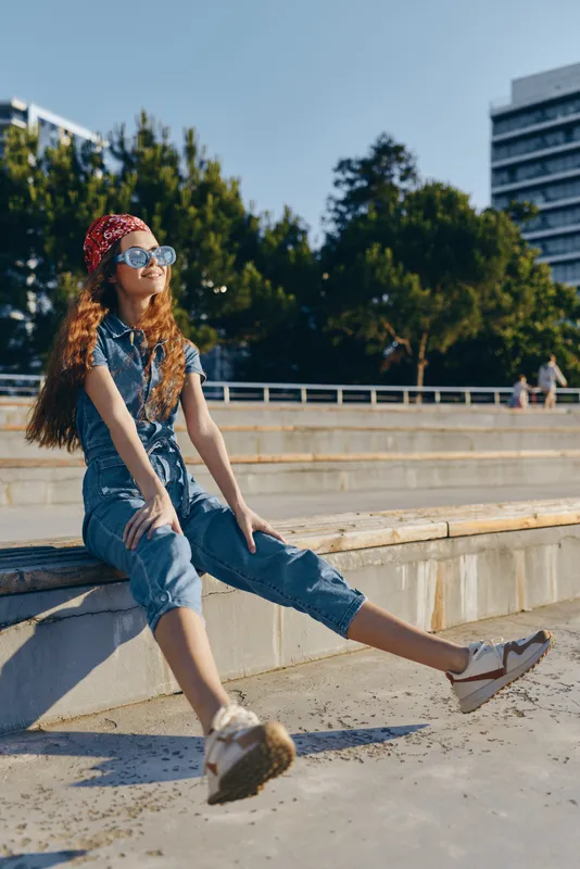 Modern boho-western denim street style woman with red bandana and sunglasses relaxing outdoors in film color sunlight, casual summer urban fashion vibe, youthful lifestyle