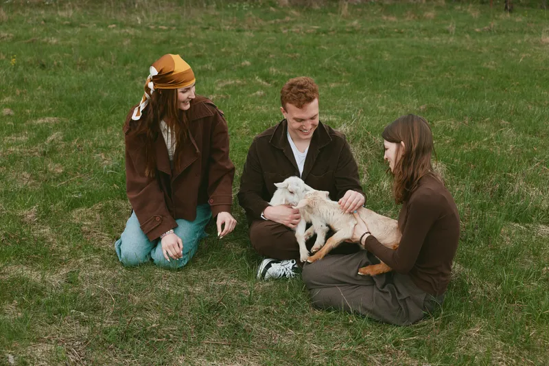 Three friends outdoors share a gentle moment with a goat kid, sitting on grass, smiling, petting, sunlit scene, casual clothing, peaceful rural vibe, natural colors, candid lifestyle
