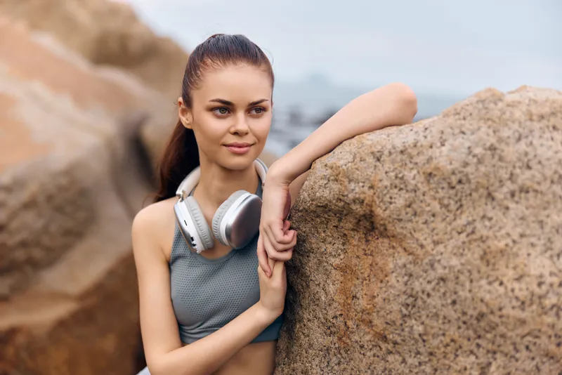 Young fitness woman with headphones at the beach, enjoying a moment of relaxation Sporty attire, natural setting, positive emotions, summer vibes, healthy lifestyle
