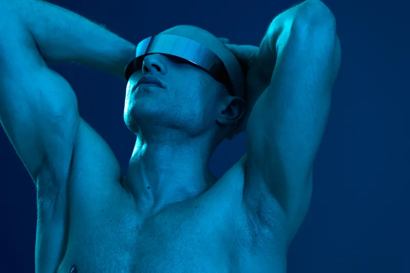 Stylish man with futuristic sunglasses posing confidently in a blue-toned environment, showcasing modern aesthetics and a bold vibe that captures attention.