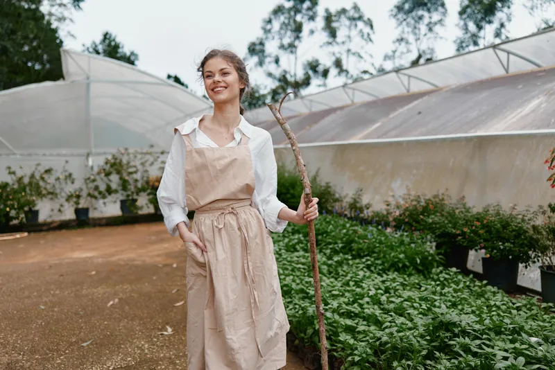 Young woman in an apron smiling joyfully in a greenhouse, holding a stick, surrounded by lush green plants and natural light, representing gardening and nature