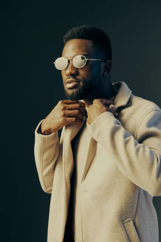Stylish African man in a beige coat and round sunglasses, exuding confidence against a dark background, showcasing modern fashion and elegance