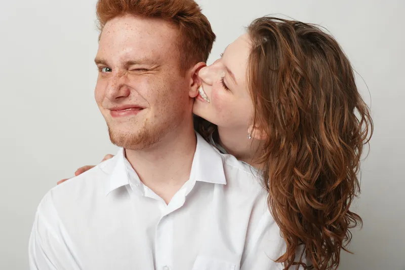 Happy young couple in white shirts share a playful moment, woman kissing man on cheek. Studio portrait with light background, concept of love, affection, and relationship joy.