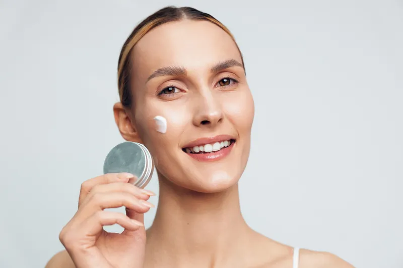 Skincare woman applying face cream with moisturizer on cheek, holding cosmetic jar near face, fresh beauty routine in daily clean minimal studio light, hygiene and self care concept