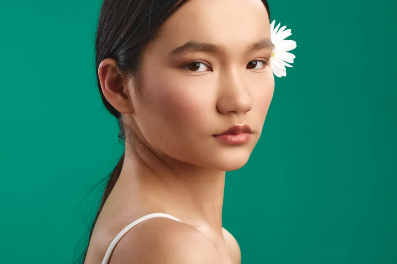 Asian woman gazes softly at the camera in a studio portrait with a vivid green background and a white flower tucked behind her ear, conveying calm elegance.