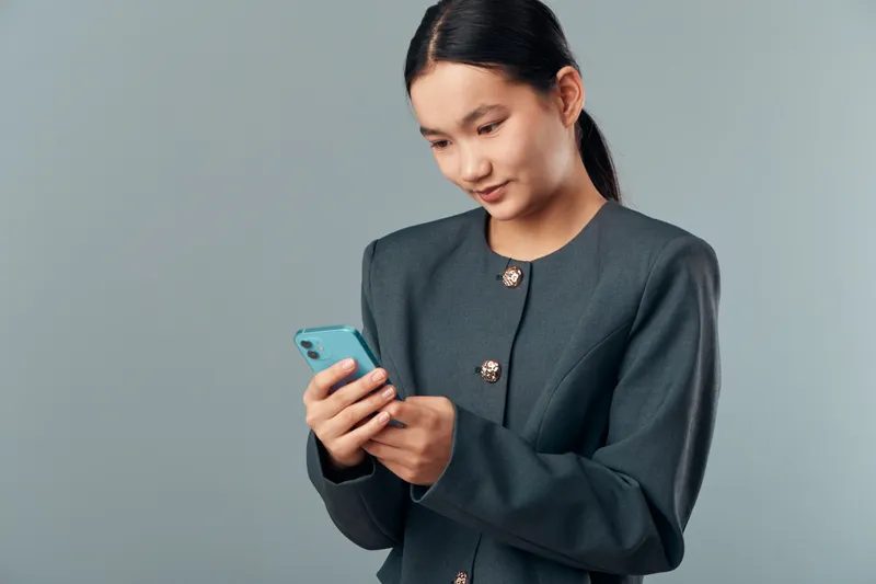 Young woman standing with smartphone in hands looking at screen for messaging or browsing with focus and calm expression on neutral background in studio.