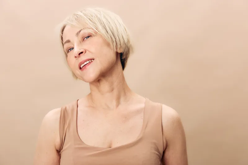Mature woman with short blonde hair smiles gently wearing beige tank top against neutral background. Concept of natural beauty, aging gracefully, confidence, and soft lighting portrait.