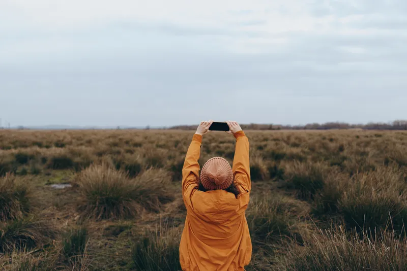Woman lifestyle portrait outdoors in an autumn field, orange hood up, hands shielding face from wind, standing among grass and tall brush, quiet solo nature mood
