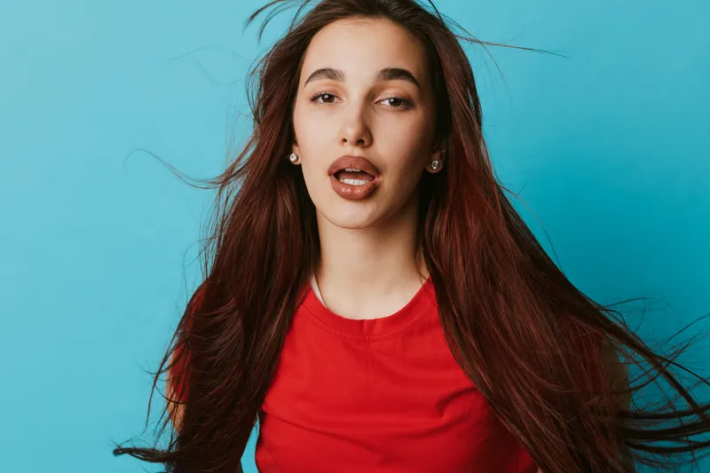 Young woman with long brown hair wearing red shirt posing with open mouth and expressive face on blue background. Portrait, beauty, casual style and natural look with loose hair movement.
