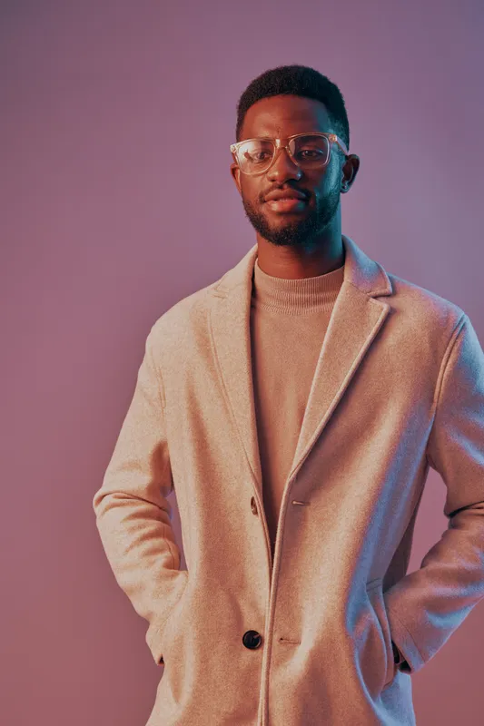 Stylish young African man in a beige coat, showcasing modern fashion with glasses against a gradient purple background, evoking confidence and sophistication