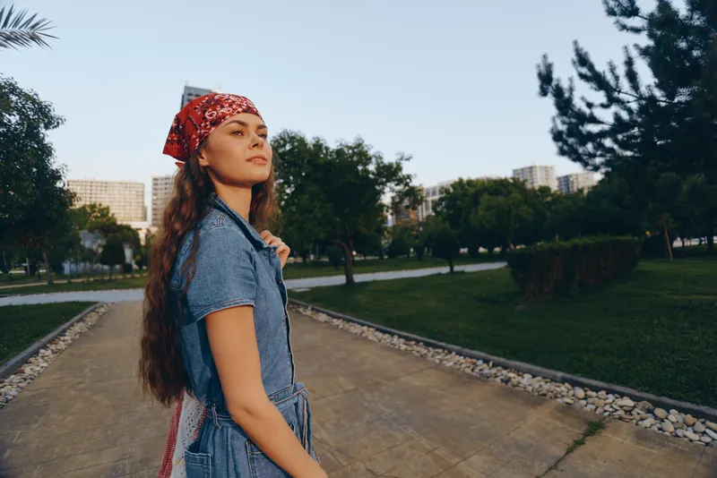 Young woman with long hair and red bandana walking outdoors in casual denim dress, enjoying peaceful urban park in warm daylight.
