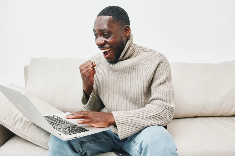 Excited Black man in cozy sweater celebrating success while working on a laptop at home, showcasing emotions of joy and accomplishment