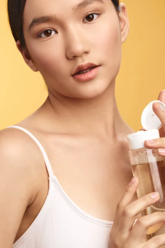 Asian woman applies lotion to her skin in a bright studio, projecting calm confidence and care with soft lighting and a minimalist, modern vibe.