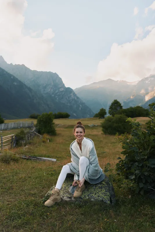 Young woman sits on a grassy hillside with distant mountains behind, smiling softly in a casual outfit, enjoying a sunlit, breezy, tranquil outdoor moment.