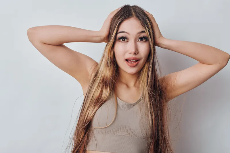 Young woman with long hair and surprised expression holding head with hands on white background, casual sleeveless top and natural makeup closeup portrait.
