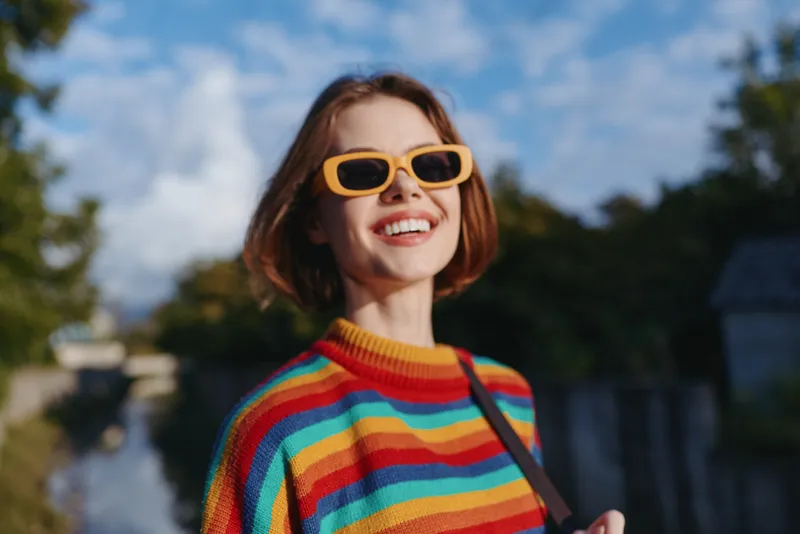 Young woman smiling woman in a rainbow sweater and yellow sunglasses in an outdoor portrait; casual fashion with short bob haircut in an urban park, walking and enjoying sunny travel lifestyle.