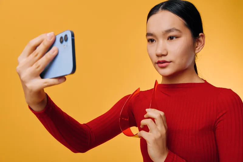 Young woman in red sweater taking selfie with smartphone on orange background. Asian girl holding phone for photo, looking at camera with focused expression in casual studio setting.