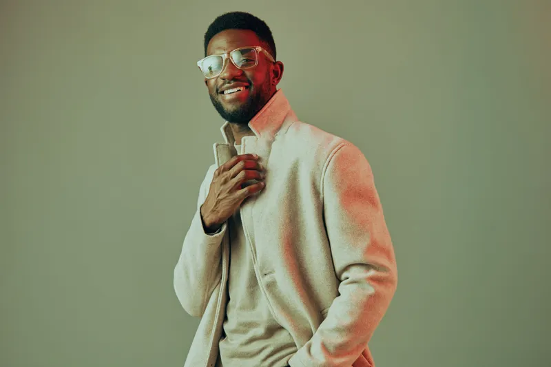 Stylish young man in glasses wearing a beige coat, smiling against a soft pastel background, showcasing modern fashion for autumn or winter