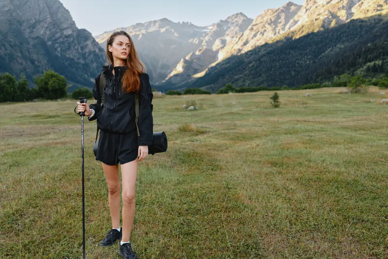 A female traveler stands in a wide alpine meadow with a trekking pole, wearing rugged outdoor gear and boots, gazing toward distant rugged peaks and endless sky.