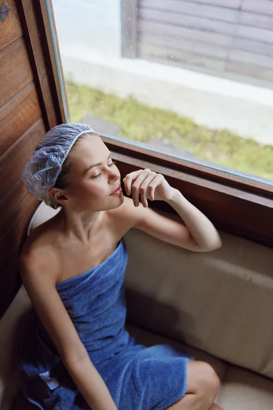 Relaxed woman wrapped in blue towel with hair cap enjoying calm moment near window, serene mood, natural light, wellness and spa concept, indoor setting.
