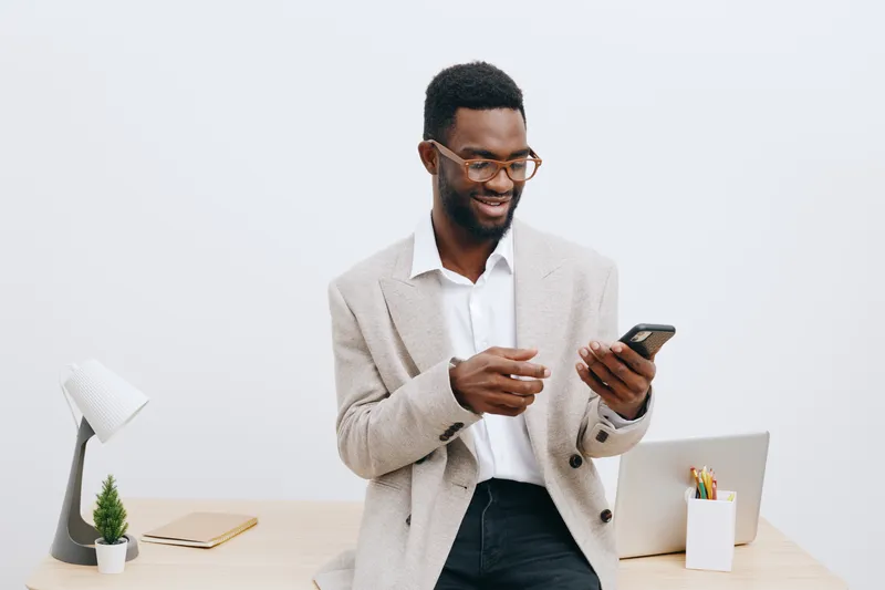 Smiling man using smartphone in office, casual beige blazer and white shirt, cheerful expression, modern workspace with laptop, plant, and stationery, bright minimalistic background. People lifestyle 