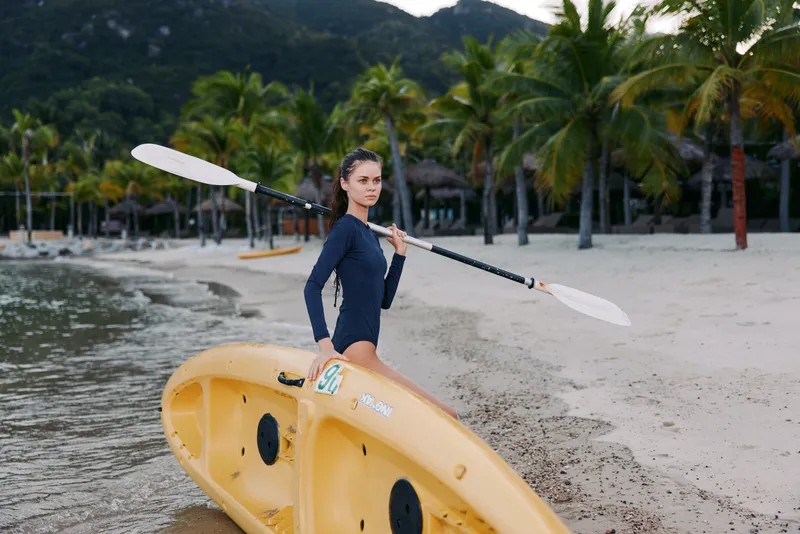 A solo female kayaker stands beside a yellow kayak on a sandy tropical beach, holding a double paddle and gazing toward the calm turquoise shoreline.