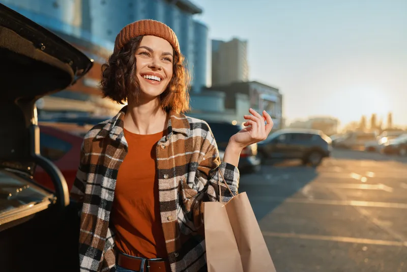 Woman smile shopping in parking at sunset by car with bag and beanie, candid lifestyle scene showing authenticity, mindful living and golden hour glow, emotional storytelling urban moment