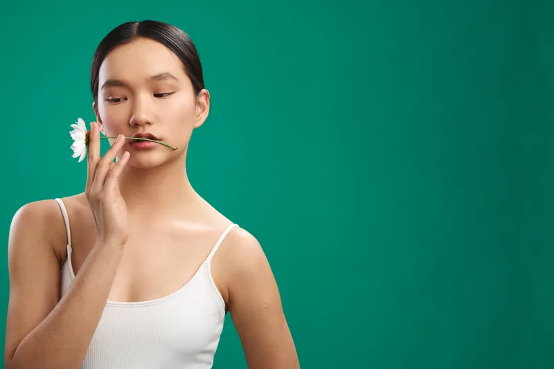 Asian woman delicately touches a flower near her lips in a simple studio setting with a teal background. She projects calmness, focus, and a graceful, minimalist mood.