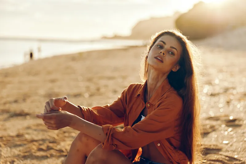 Young woman sitting on beach at sunset, stretching her arms toward the sea, warm golden hour sunlight, outdoor sea waves and sparkles in background, freedom and joy for relaxed mood
