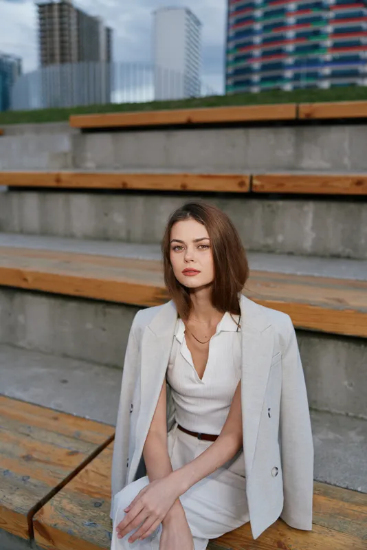 Young businesswoman in white suit sitting on urban wooden steps with thoughtful expression, modern cityscape background
