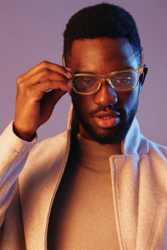 Young Black man adjusting stylish glasses, dressed in a modern light gray coat, displaying a thoughtful expression under a vibrant purple backdrop