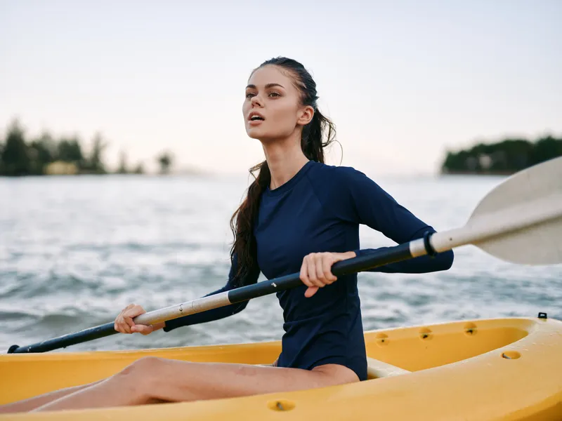 A focused kayaker paddles an orange kayak along calm water at golden hour, wearing a dark blue athletic suit and gripping a double?blade paddle with confident, steady strokes.