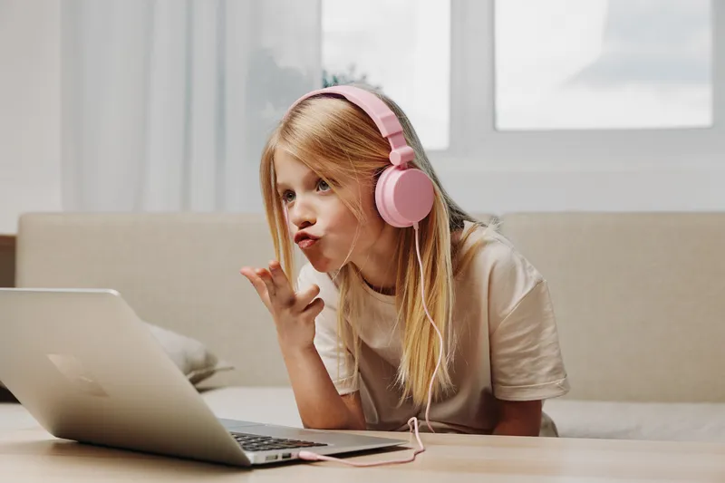 Blond girl with pink headphones sends a kiss to laptop screen while sitting on couch in bright room. Concept of online communication and digital interaction for children.