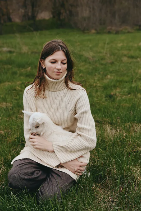 Calm woman outdoors, seated on grass, holding a goat kid, cozy cream sweater, soft natural light, tranquil expression, nature background, relaxed lifestyle moment, tender connection