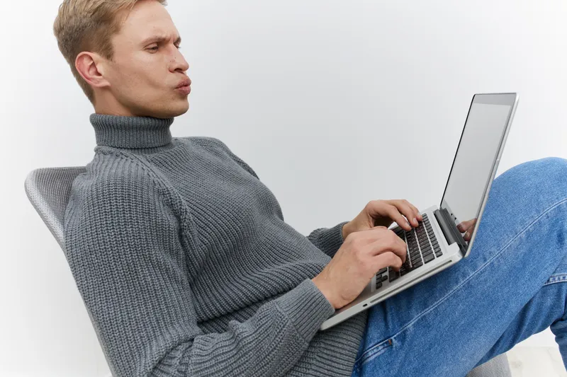 Man working on laptop computer wearing gray turtleneck sweater and blue jeans sitting on white background. Casual businessman using technology. People lifestyle concept.