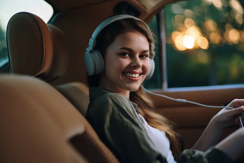 Smiling young woman with headphones enjoying music in a car during sunset, showcasing joy and relaxation. Perfect for themes of youth, happiness, and leisure.
